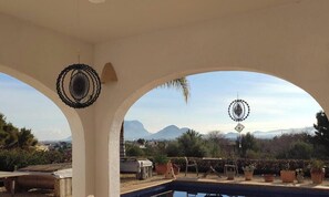 A pool area with a view of mountains, a covered patio with arches, and decorative hanging items.