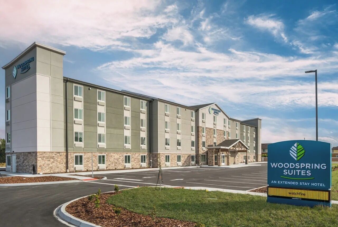 Um hotel com vários andares, fachada de pedra, entrada central e uma placa indicando ser um hotel Woodspring Suites para estadias prolongadas.