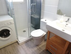 A bathroom with a glass-enclosed shower, a washing machine, a toilet, and a wooden cabinet. - Beachfront Apartment in Denia (Denia)