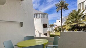 A small outdoor patio with a round table and four chairs, a white building with a curved balcony, and a palm tree in the background.