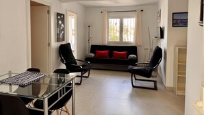 A living room with a glass table, two black chairs, a black sofa with red cushions, a TV, and a window with curtains.
