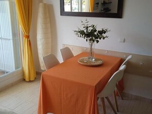 A dining area with a table covered in orange, surrounded by white chairs, a vase with greenery, and a window with yellow curtains.
