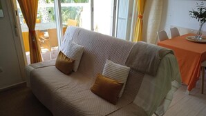 Et værelse med en beige sofa, to puder, et bord med orange dug og en plante på bordet.