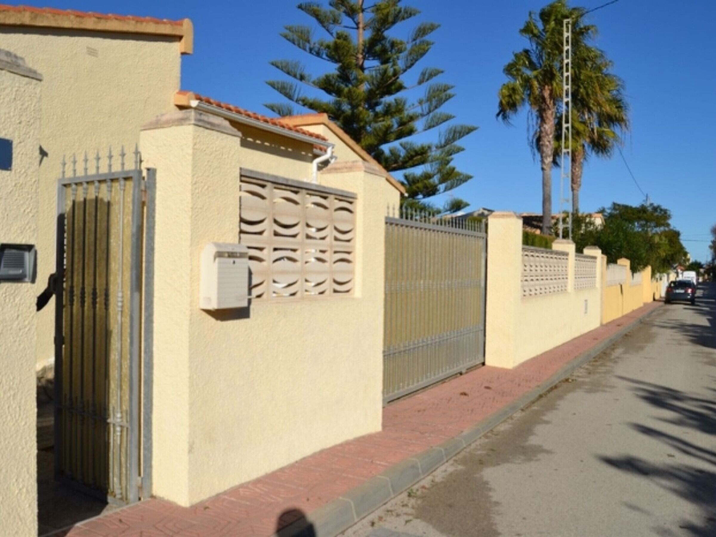 A residential street with a beige house, a metal gate, and a palm tree.