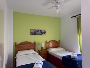 A hotel room with two beds, a ceiling fan, a nightstand, a lamp, and a painting on the wall.
