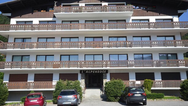 Un hotel a più piani con balconi, un cartello con su scritto "L'ALPENROSE" e auto parcheggiate di fronte.