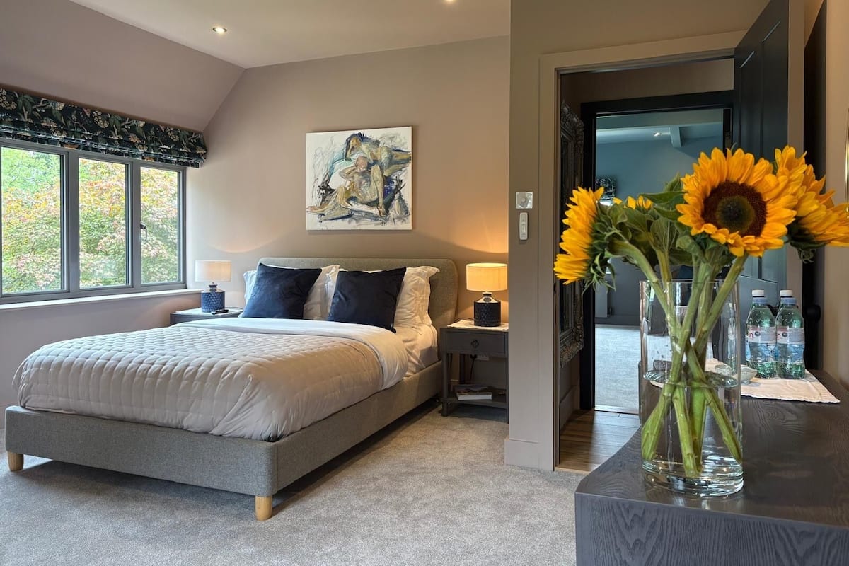 A modern bedroom with a large bed, a nightstand, a painting on the wall, and a vase of sunflowers on a table.