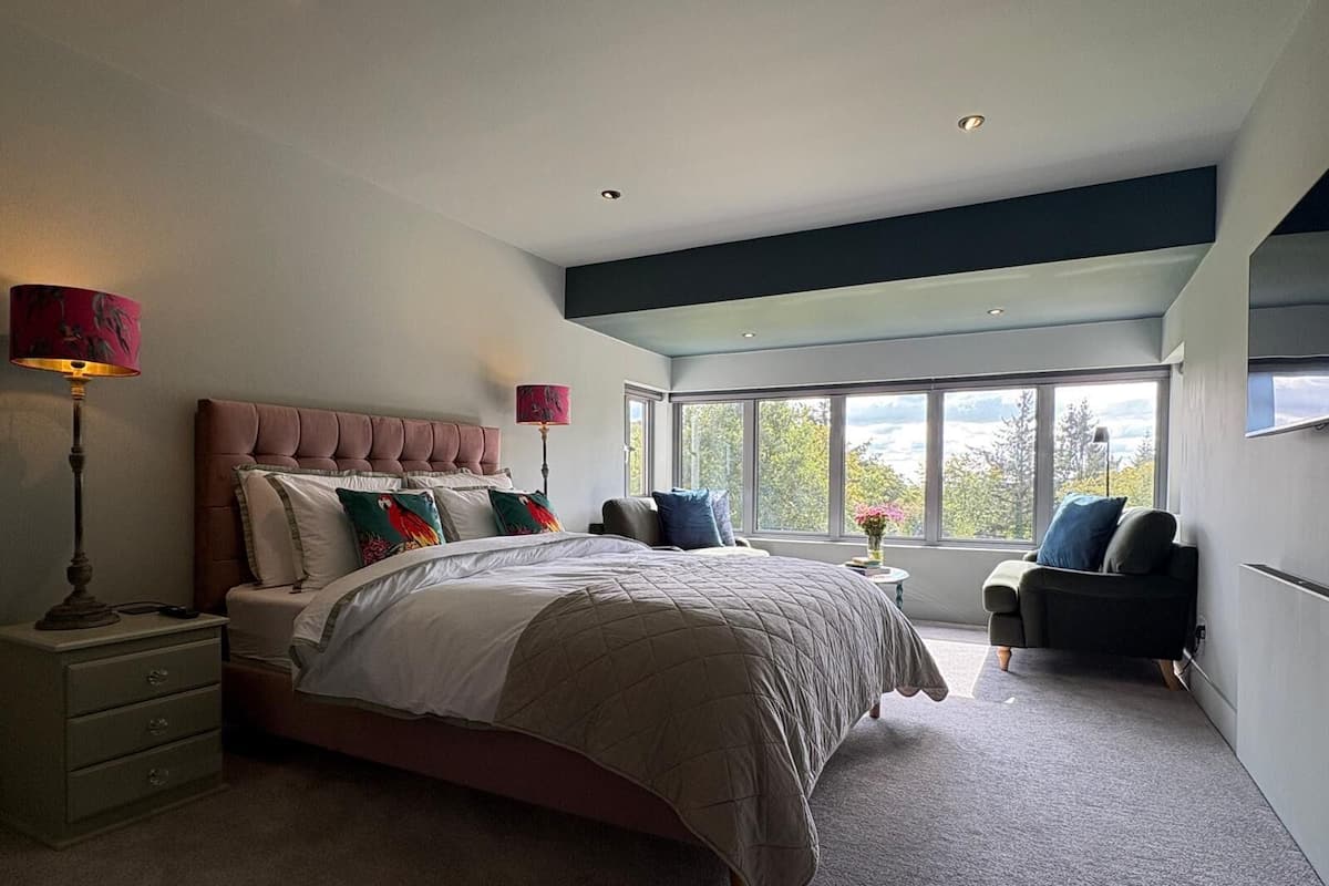 A modern bedroom with a large bed, bedside lamps, a window with a view, and a small table with a vase.