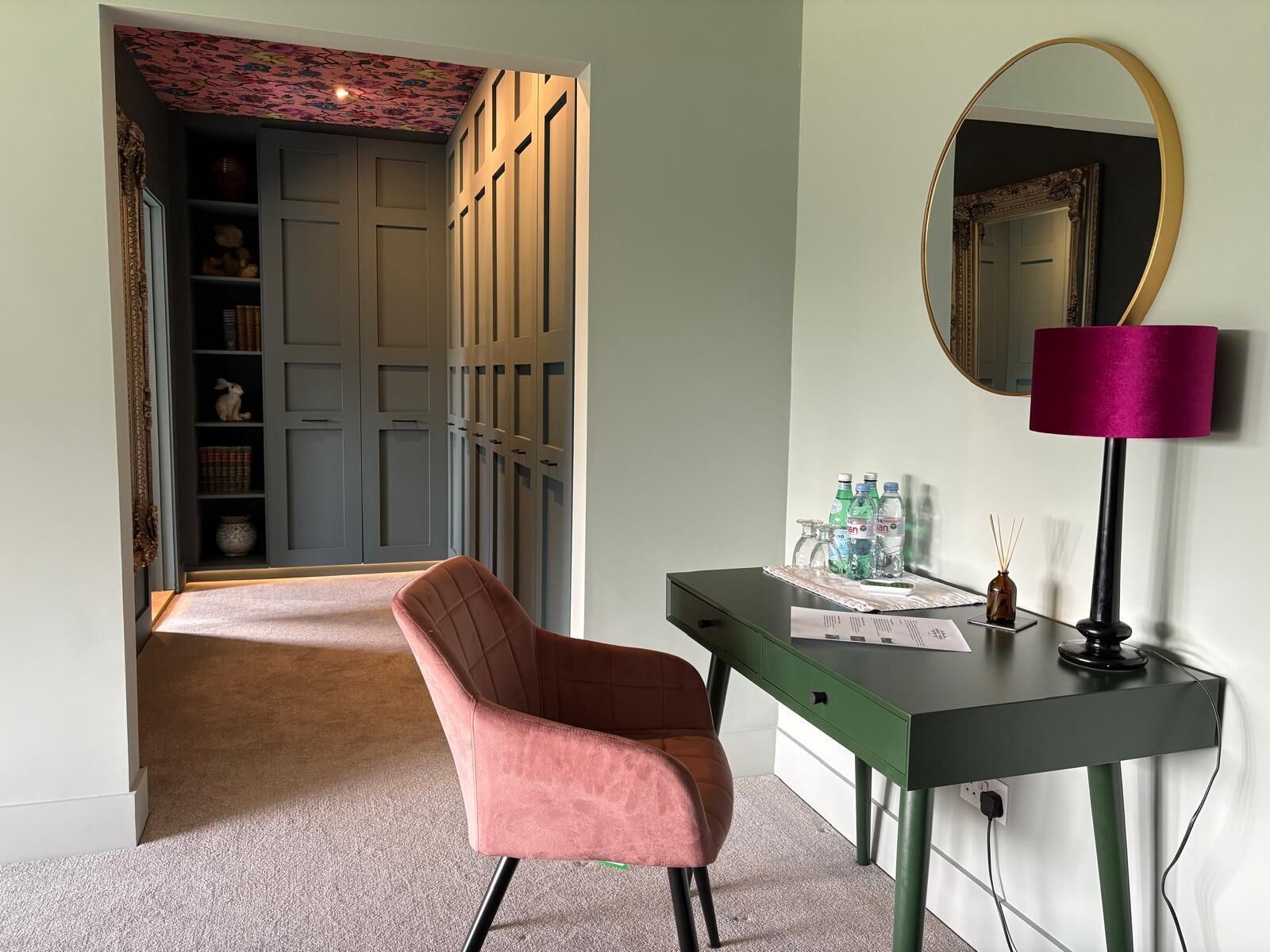 A room with a desk, chair, mirror, and a wardrobe with shelves.