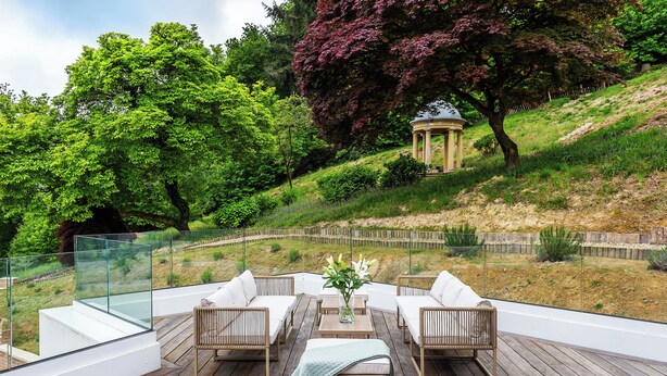 Um deck de madeira com mobiliário externo, guarda-corpo de vidro e vista para um jardim paisagístico com árvores e um gazebo.