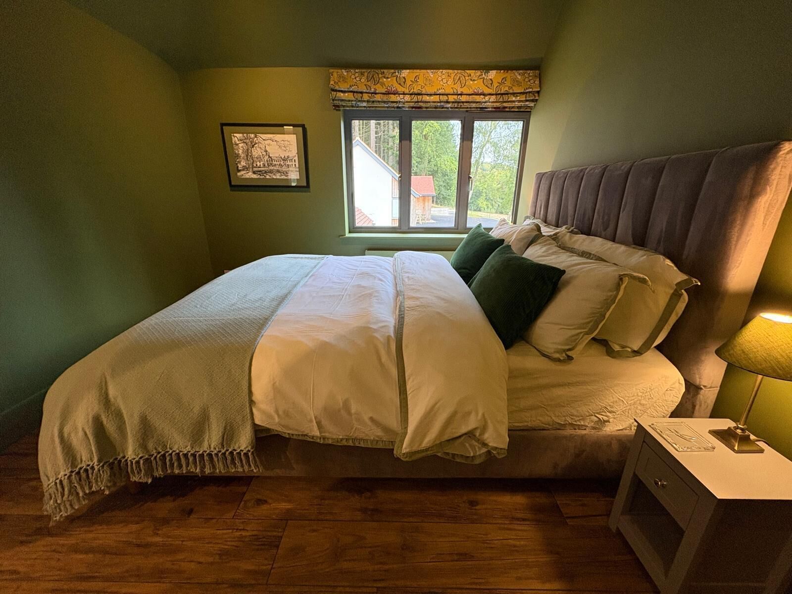 A bedroom with a large bed, a nightstand, a lamp, a window with a view of trees, and a framed picture on the wall.