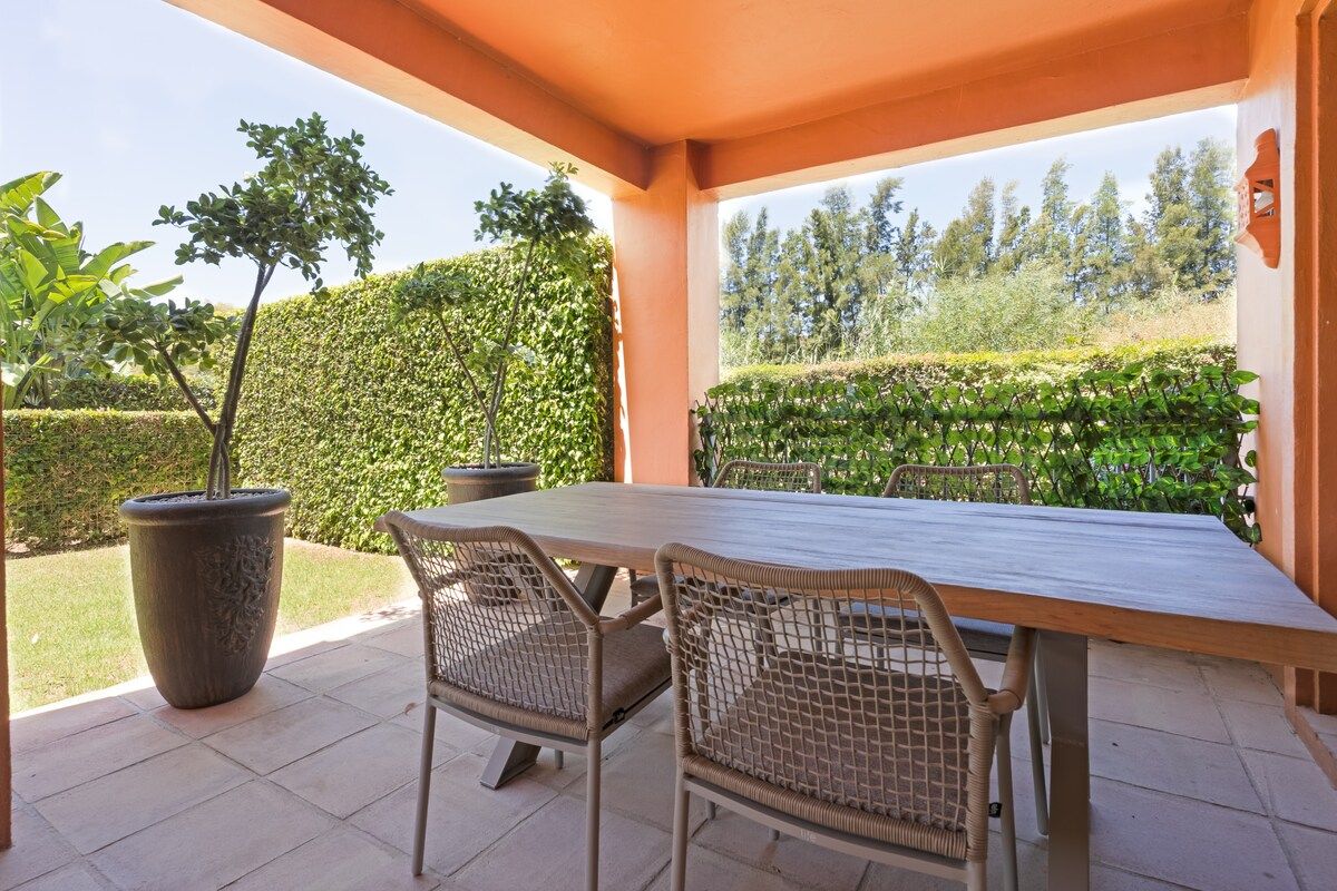 En udendørs terrasse med et bord og stole, omgivet af grøn vegetation og udsigt til træer.