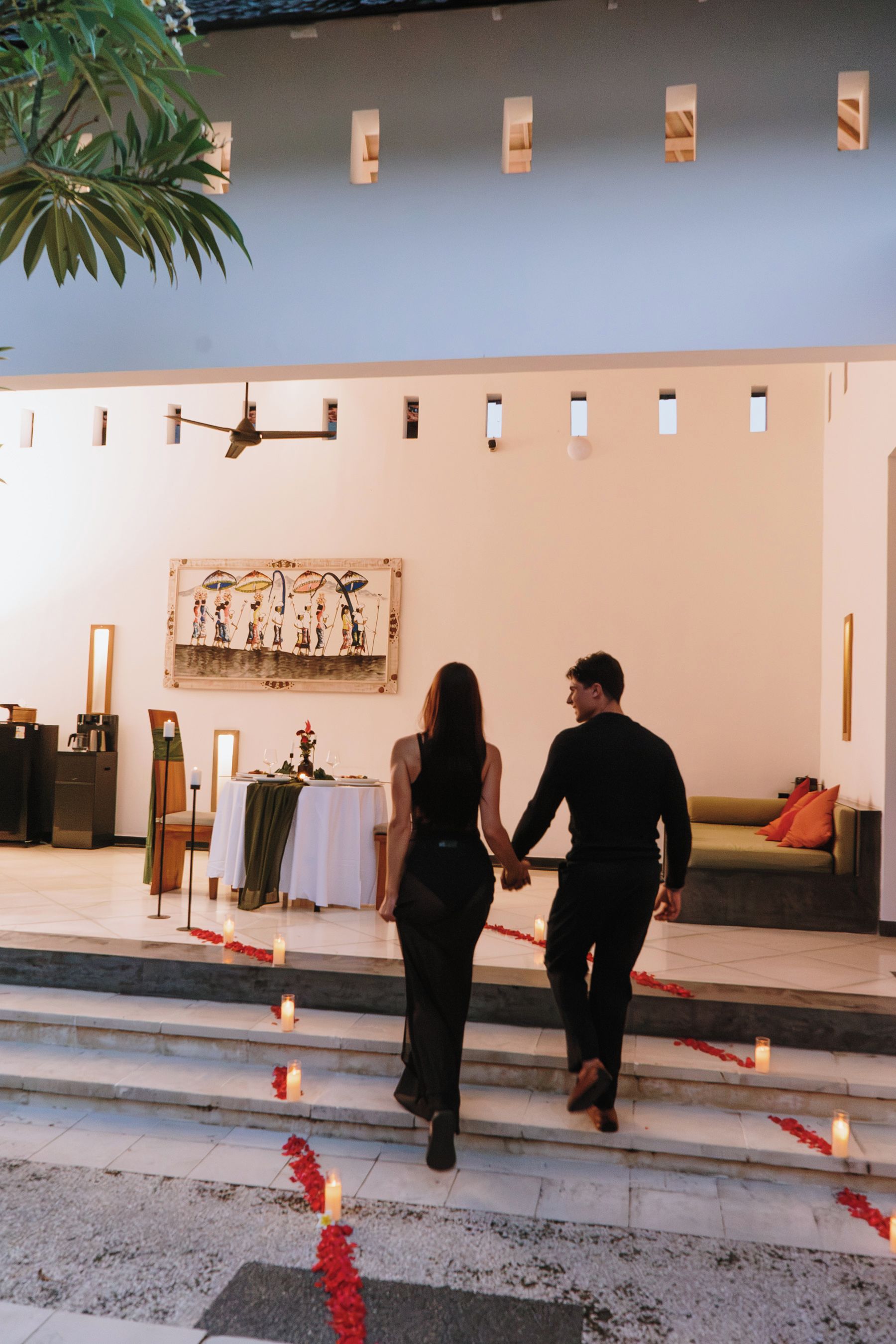 Un couple descend un escalier décoré de bougies et de pétales rouges, qui mène à une salle à manger où une table est dressée pour deux personnes.