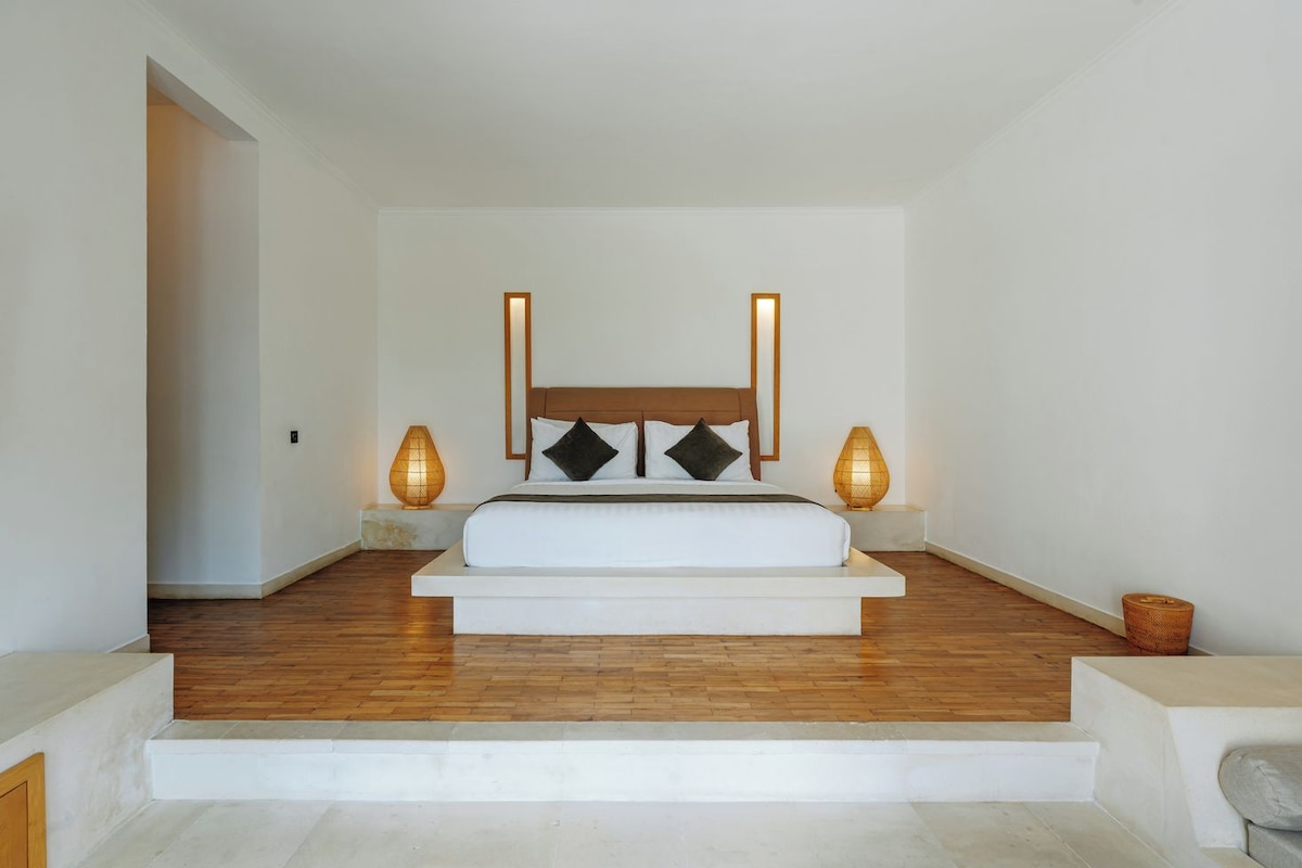 A bedroom with a wooden floor, a white bed, two bedside lamps, and a door leading to another room.