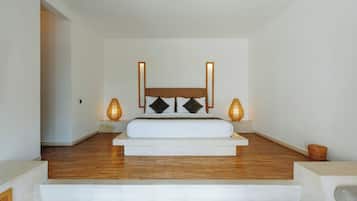 Quarto com piso de madeira, uma cama branca, duas luminárias de cabeceira e uma porta que dá para outro cômodo.