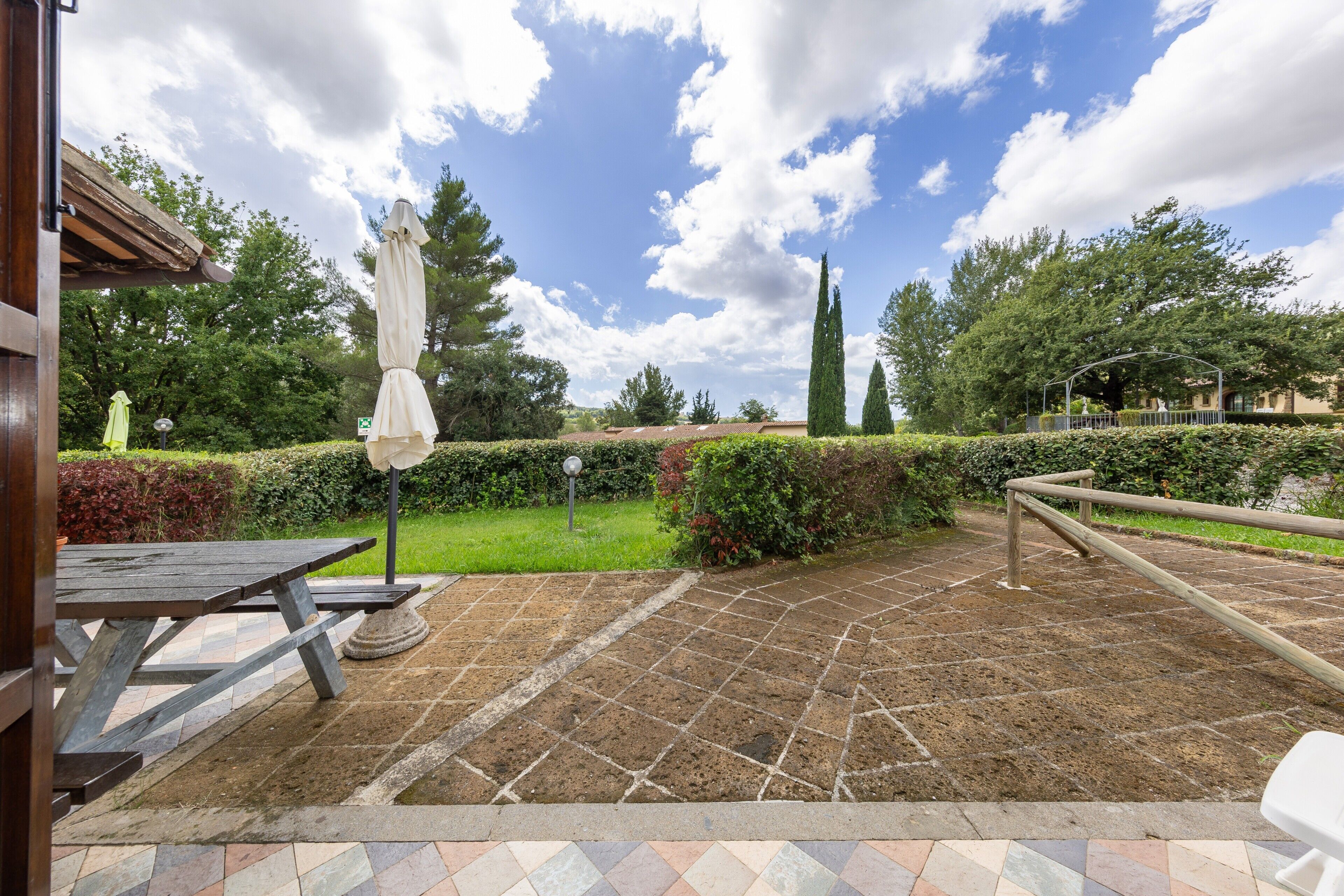 A patio area with a wooden table, a white umbrella, and a stone-tiled walkway.