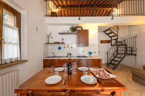 A dining area with a wooden table set for four, a kitchen in the background, and a staircase leading to an upper level.