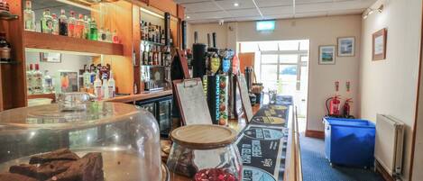 Een bar met verschillende drankflessen, een toonbank met een glazen pot en een bord, en een blauwe prullenbak.