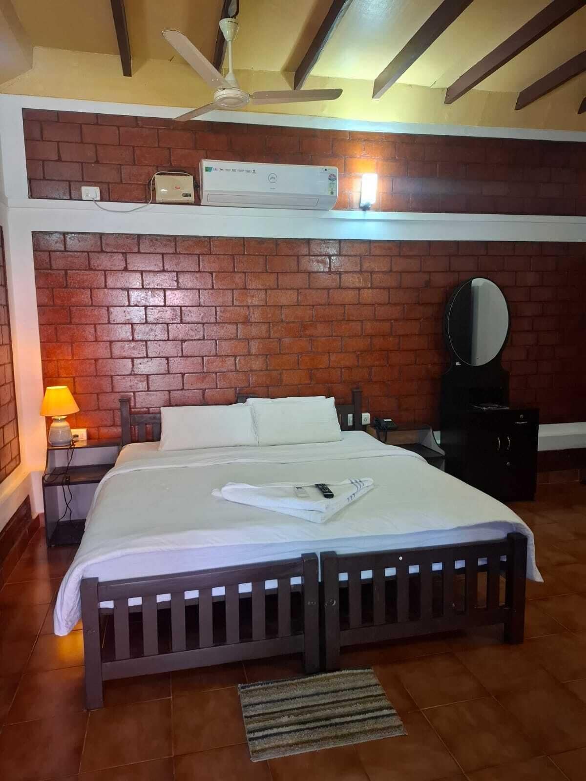 Een hotelkamer met een bakstenen muur, een bed met witte lakens, een plafondventilator, een klein tafeltje met een lamp en een spiegel aan de wand.
