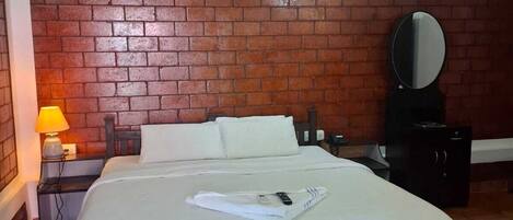 Habitación de hotel con pared de ladrillo, cama con ropa blanca, ventilador de techo, pequeña mesa con lámpara y espejo en la pared.