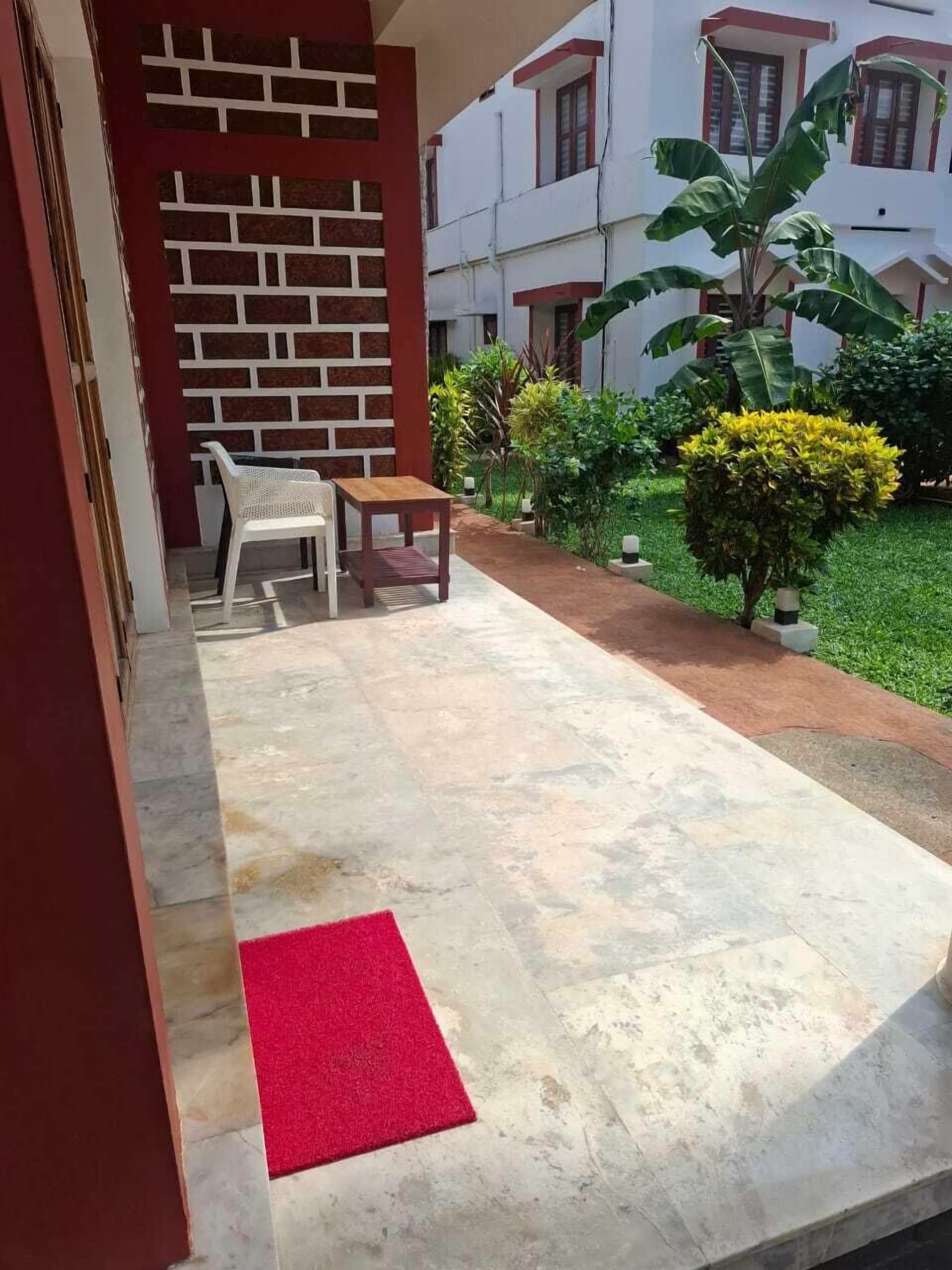 A courtyard with a red mat, a white chair, a wooden table, and a brick wall with a window.