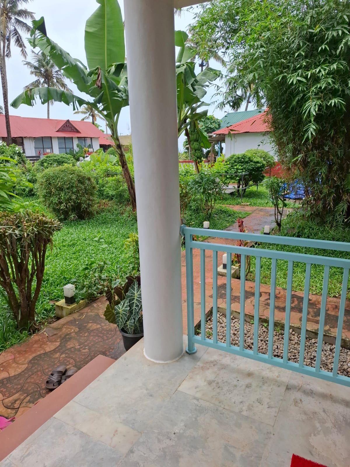 A garden area with a white column, a blue railing, and a pathway leading to a building with a red roof.