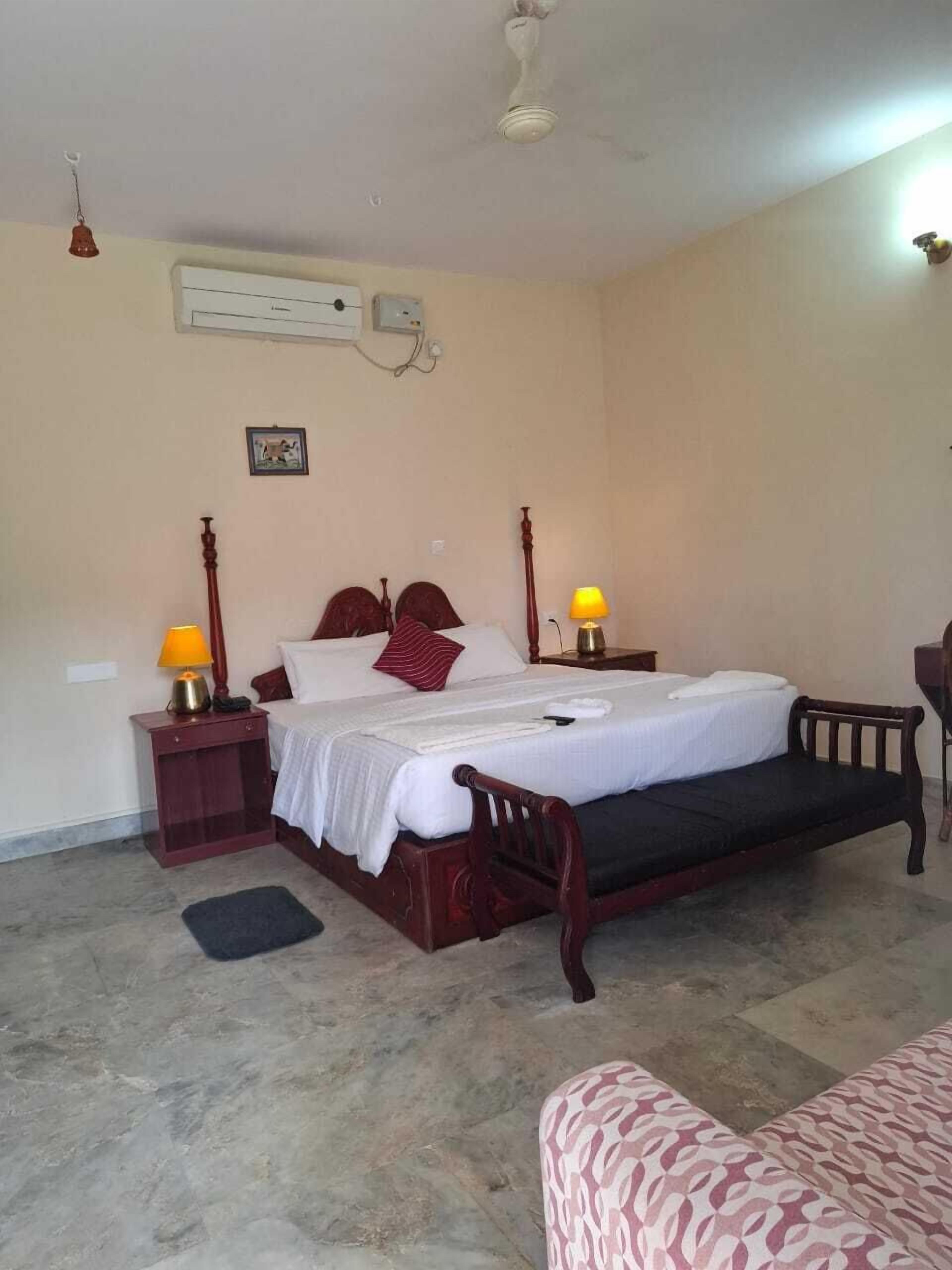 A bedroom with a bed, bedside lamps, a ceiling fan, an air conditioning unit, and a patterned sofa.