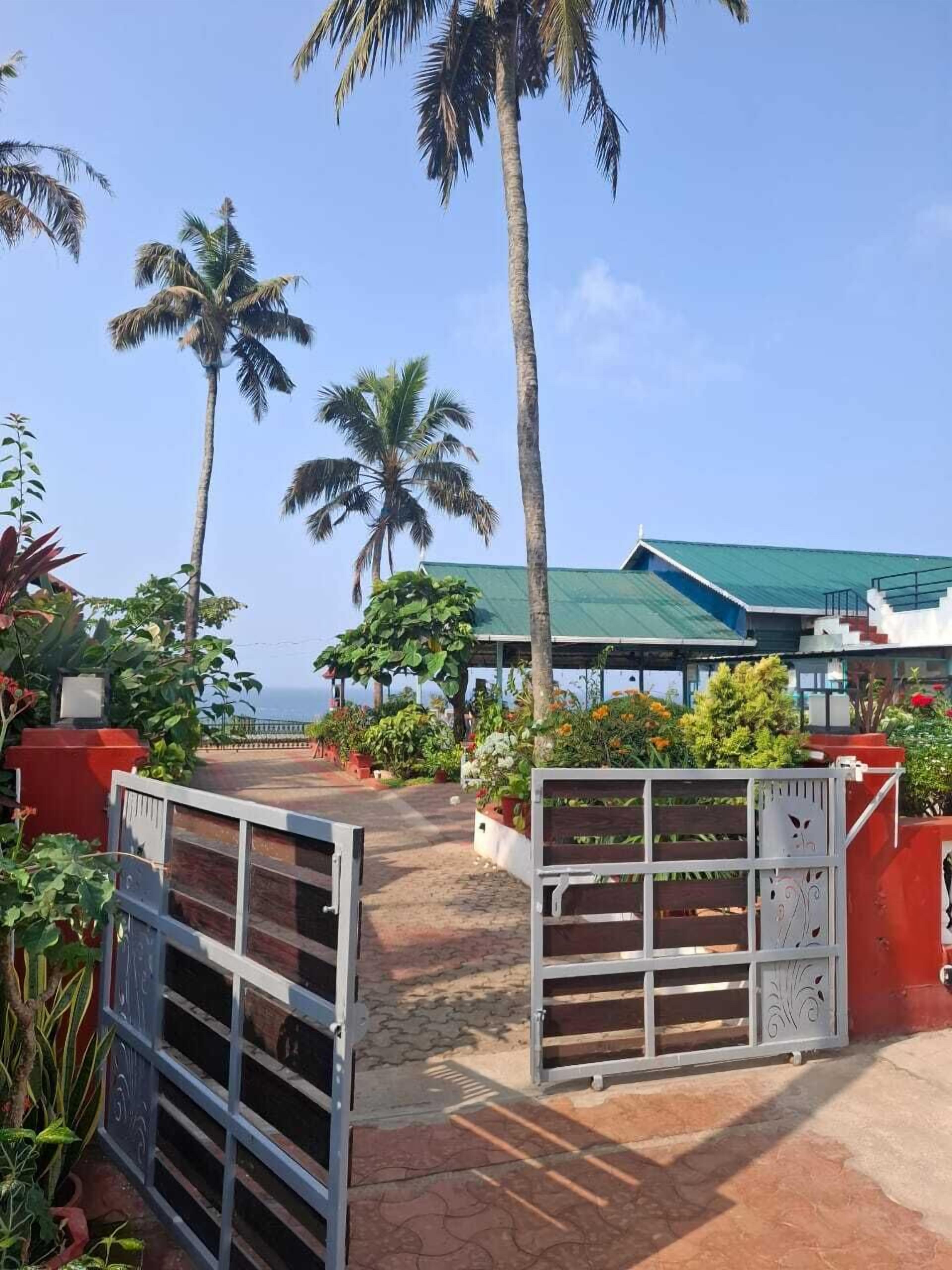 A gated entrance with palm trees, a covered area with a green roof, and a view of the sea.