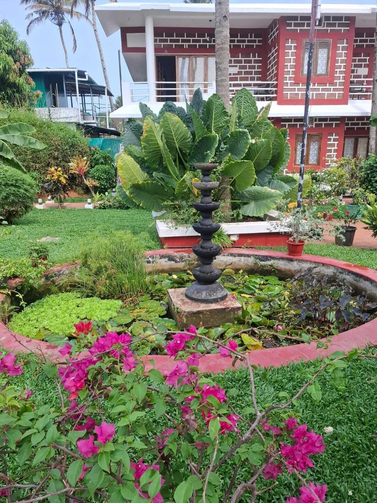 A garden with a fountain, lush greenery, and a building with a red and white facade in the background.
