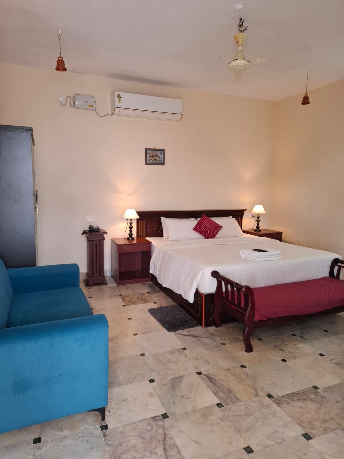 A hotel room with a bed, bedside lamps, a blue armchair, a marble floor, and an air conditioning unit.