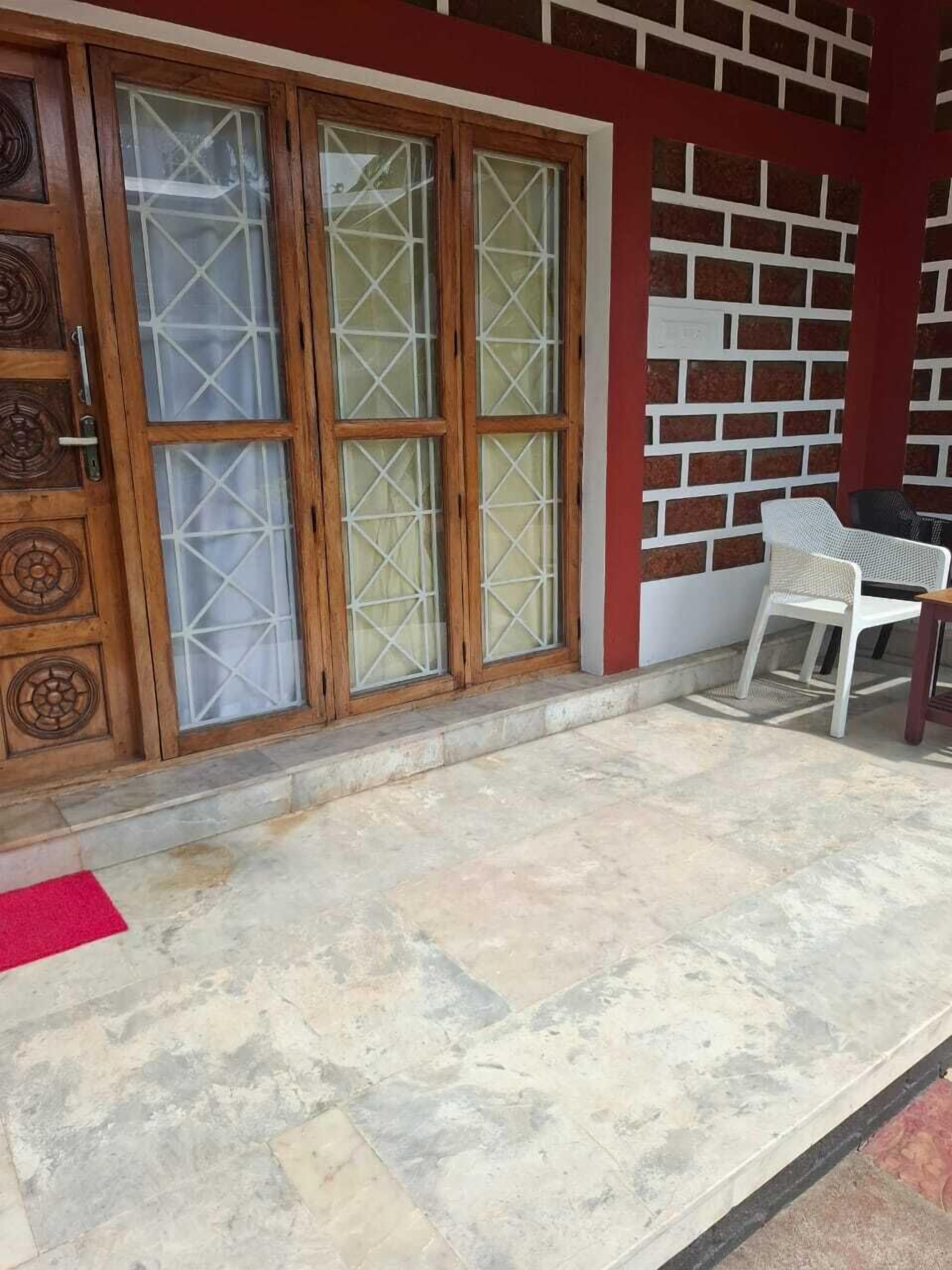 A building entrance with a wooden door with glass panels, a red brick wall, and a small outdoor seating area with plastic chairs.
