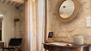 Une pièce rustique avec un mur en pierre, un miroir en osier, une table en bois sur laquelle se trouvent un livre et une carafe en céramique, et une chaise noire.