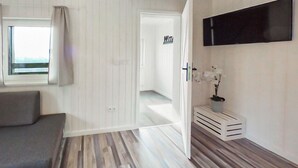 Een moderne kamer met een grijze bank, een houten vloer, een witte deur met een 'WTA' bord en een wandtv, plus een potorkidee.