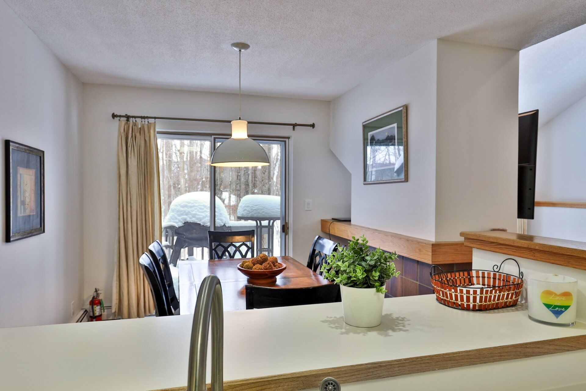 A modern kitchen with a dining area, a large window with a view of snow-covered trees, and a hanging light fixture.
