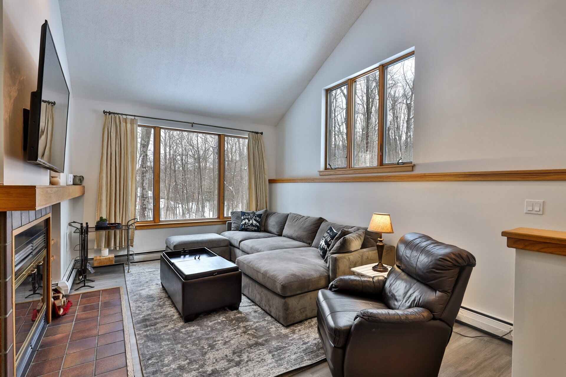 A modern living room with a sectional sofa, a leather armchair, a coffee table, and a TV mounted on the wall. There are two windows providing views of trees and a fireplace with a mantel.