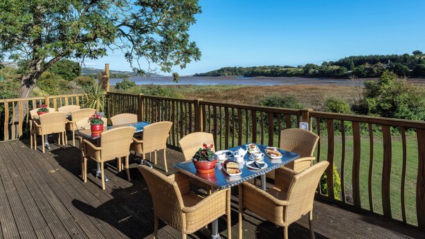 A deck with wicker furniture, a table set for dining, and a view of a body of water and hills. - Avocet (Newton Abbot)
