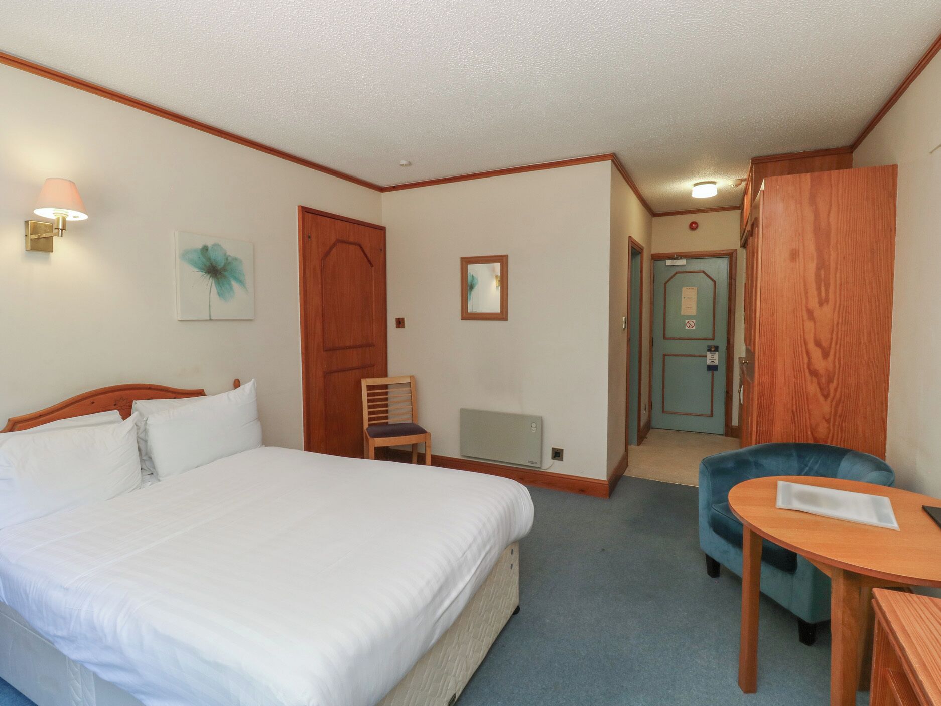A hotel room with a bed, a desk, a chair, a radiator, and a door leading to another room.