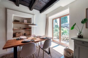 A dining area with a wooden table, chairs, and a hanging light fixture. There are shelves above the table holding baskets and books. A large window provides a view of the outdoors. - Casa Mina (Menaggio)