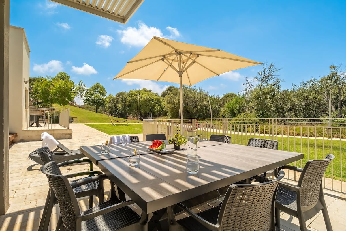 An outdoor patio with a table and chairs, a large umbrella, and a view of a grassy area and trees.