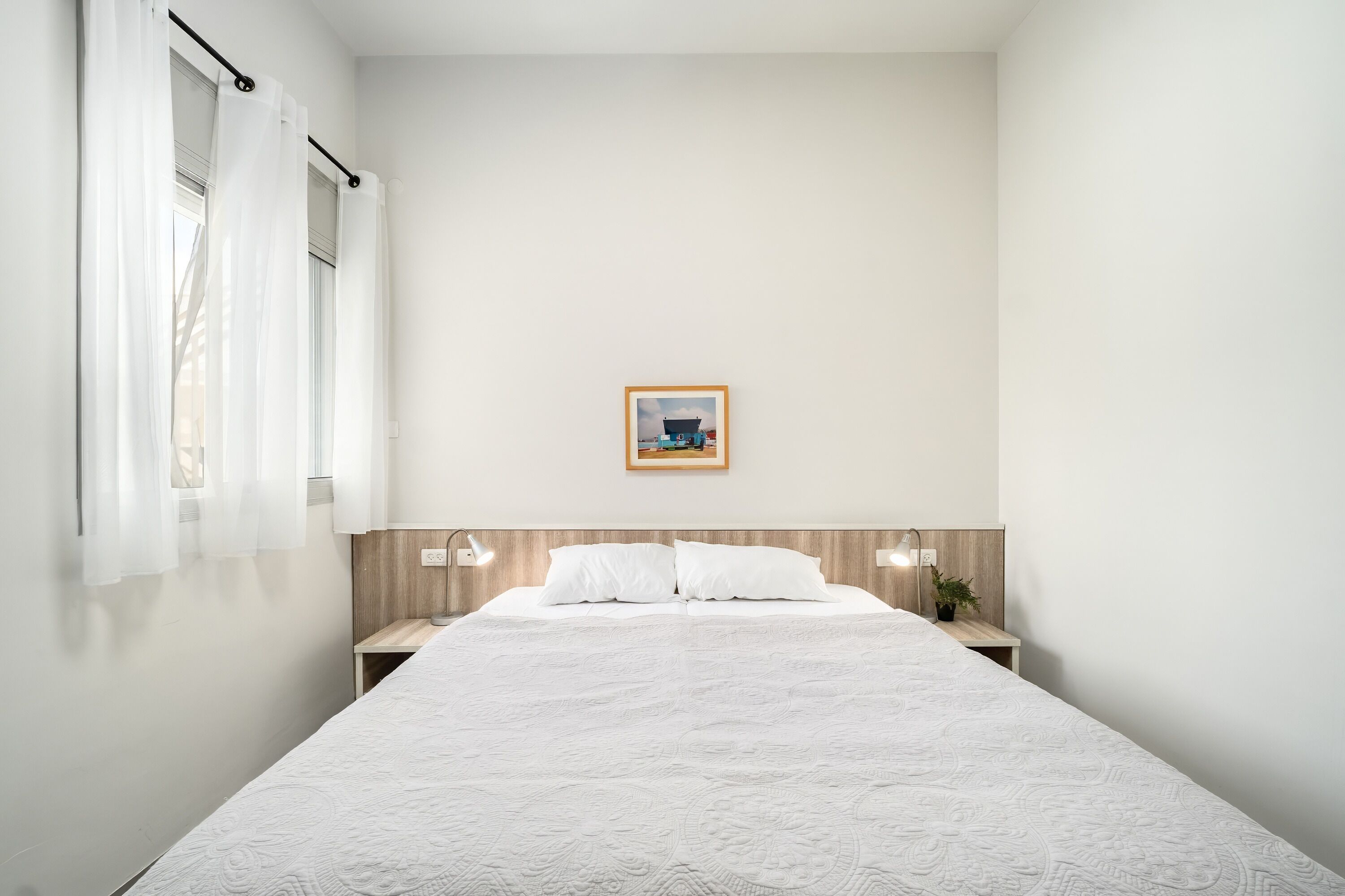 A minimalist bedroom with a large bed, two bedside tables, a nightstand, a framed picture on the wall, and sheer curtains on the window.
