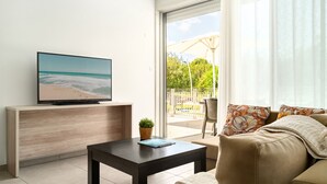 A modern living room with a flat-screen TV on a wooden console, a coffee table, a sofa with patterned cushions, and a view of a balcony with outdoor furniture.
