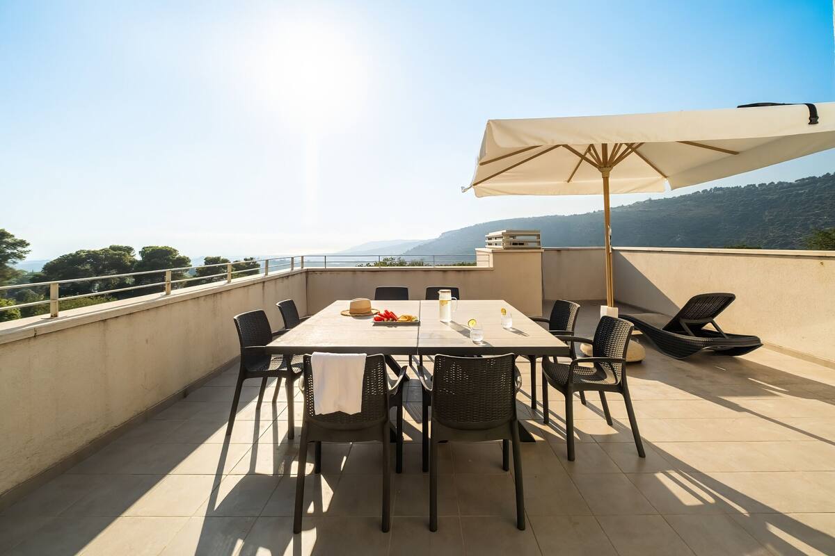 A rooftop terrace with a table set for four, surrounded by chairs, a large umbrella, and a scenic view of the sea and hills.