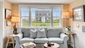 Ein gemütliches Wohnzimmer mit grauem Sofa, einem kleinen Tisch und Blick auf ein Haus durch große Fenster.