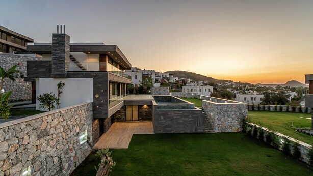 Une maison moderne avec une façade en pierre, une piscine sur le toit et une vue sur un quartier résidentiel au coucher du soleil.