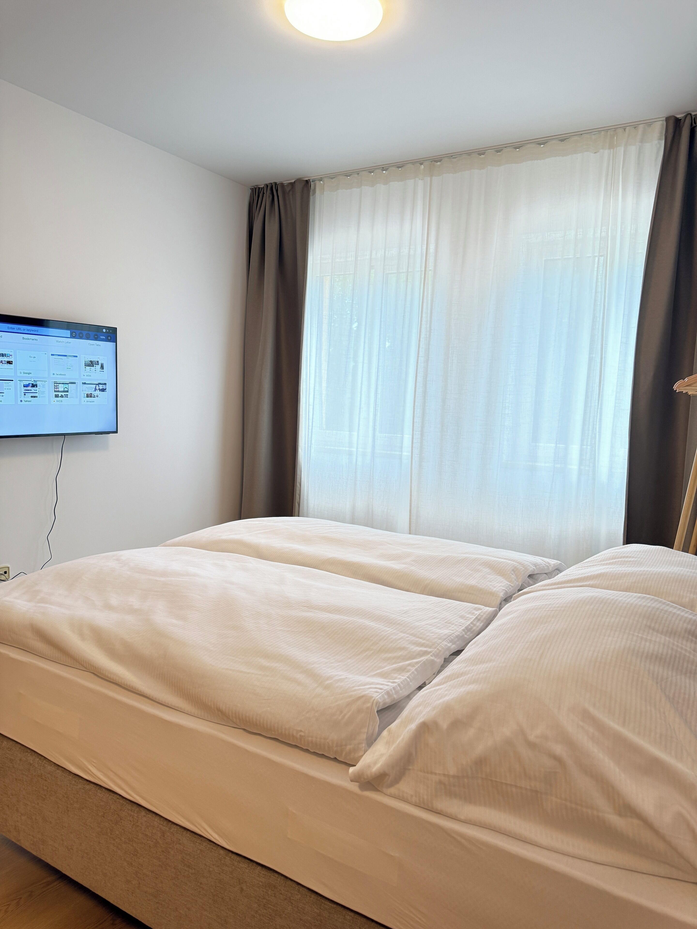A hotel room with a bed, a flat-screen TV mounted on the wall, and a window with sheer curtains.
