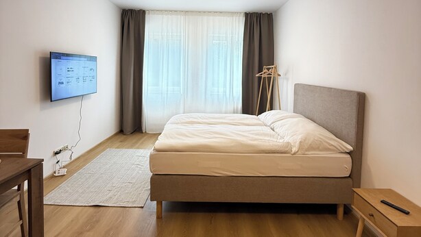 Une chambre à coucher avec un lit, un téléviseur fixé au mur, un bureau en bois et une lampe sur pied.