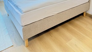 Un lit bien fait, avec une tête de lit grise, dans une pièce dotée d’un plancher en bois et d’une fenêtre ornée de rideaux légers.