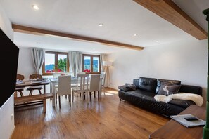 Ein modernes Wohnzimmer mit Holzboden, einer schwarzen Ledersofa, einem Esstisch mit Stühlen und Blick durch große Fenster.