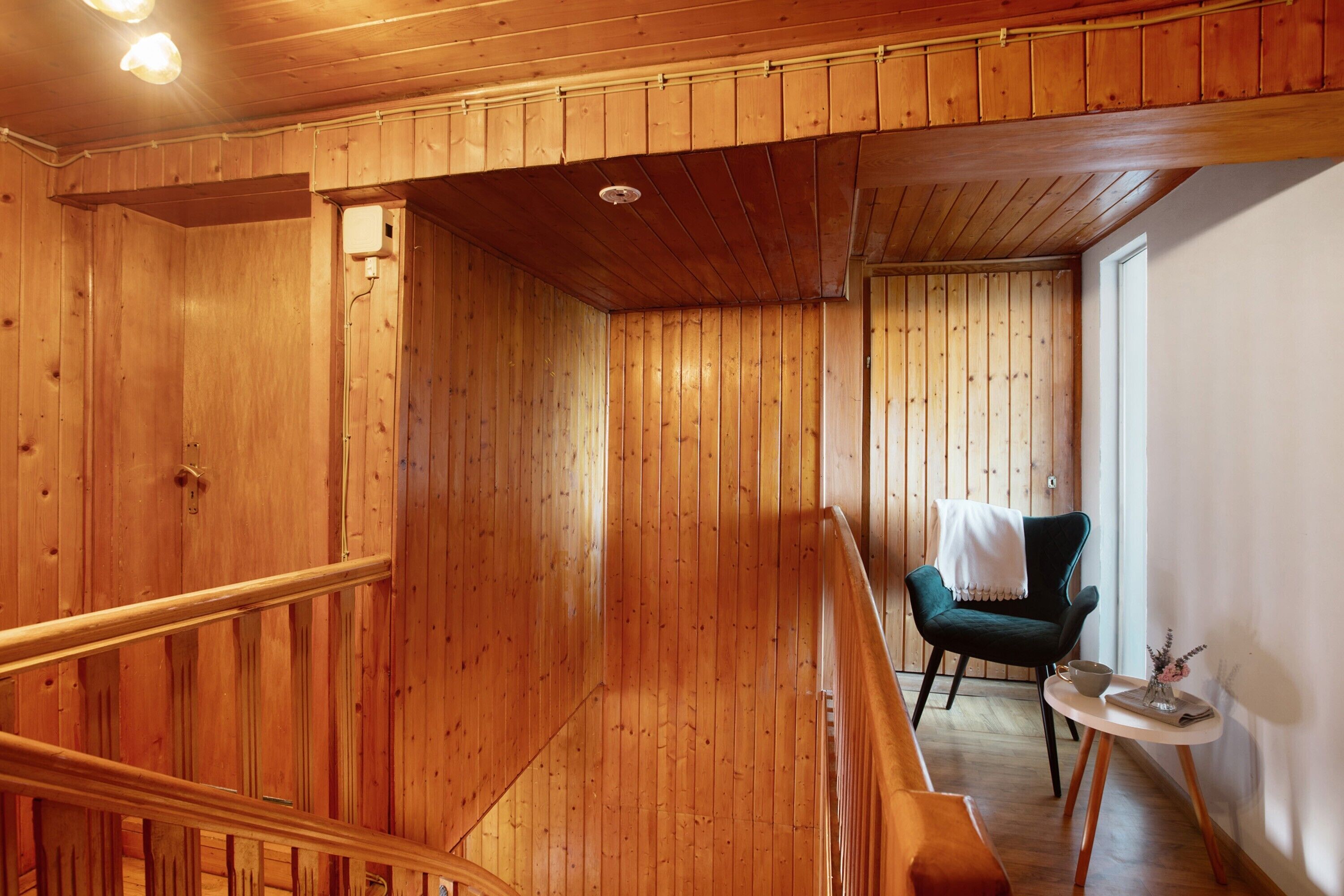 A wooden interior with a staircase, a green chair, a small table with a vase, and a window.