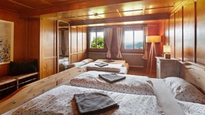 A room with wooden walls and ceiling, two beds with floral bedding, a bench, a lamp, and a window with a view of greenery.